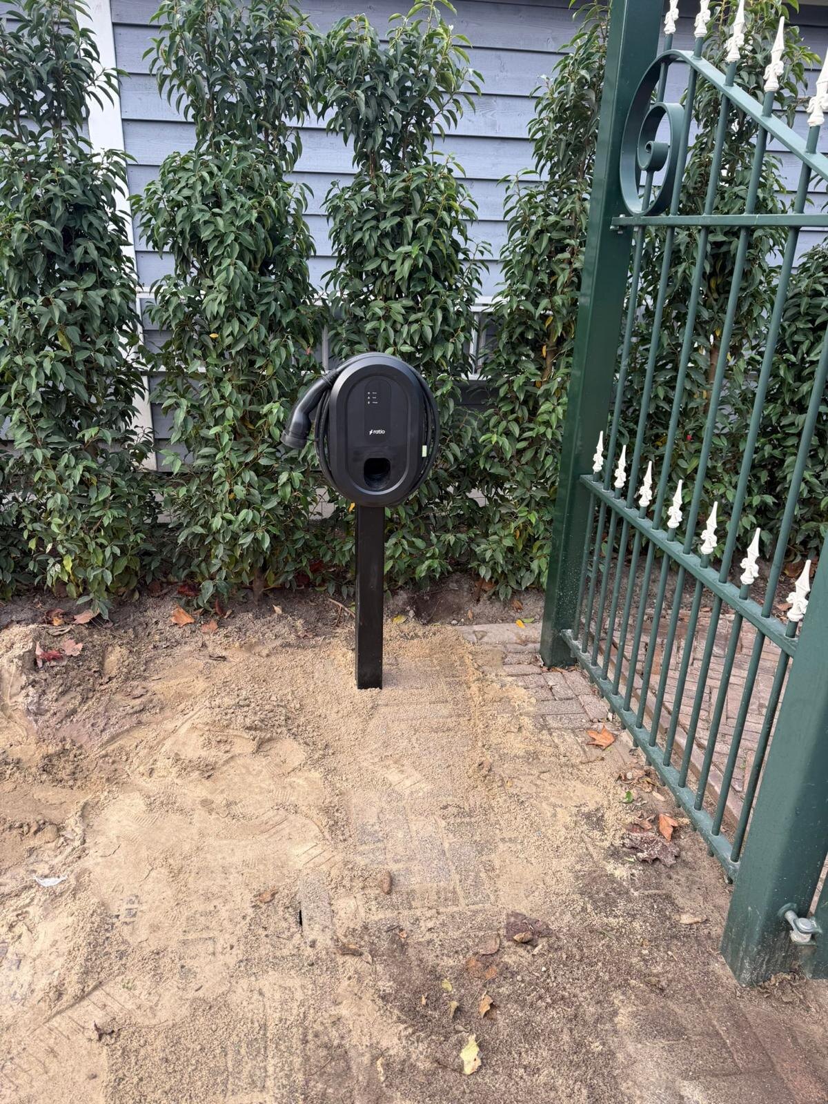 Vrijstaande laadpaal op een statief geplaatst in een tuin naast een poort