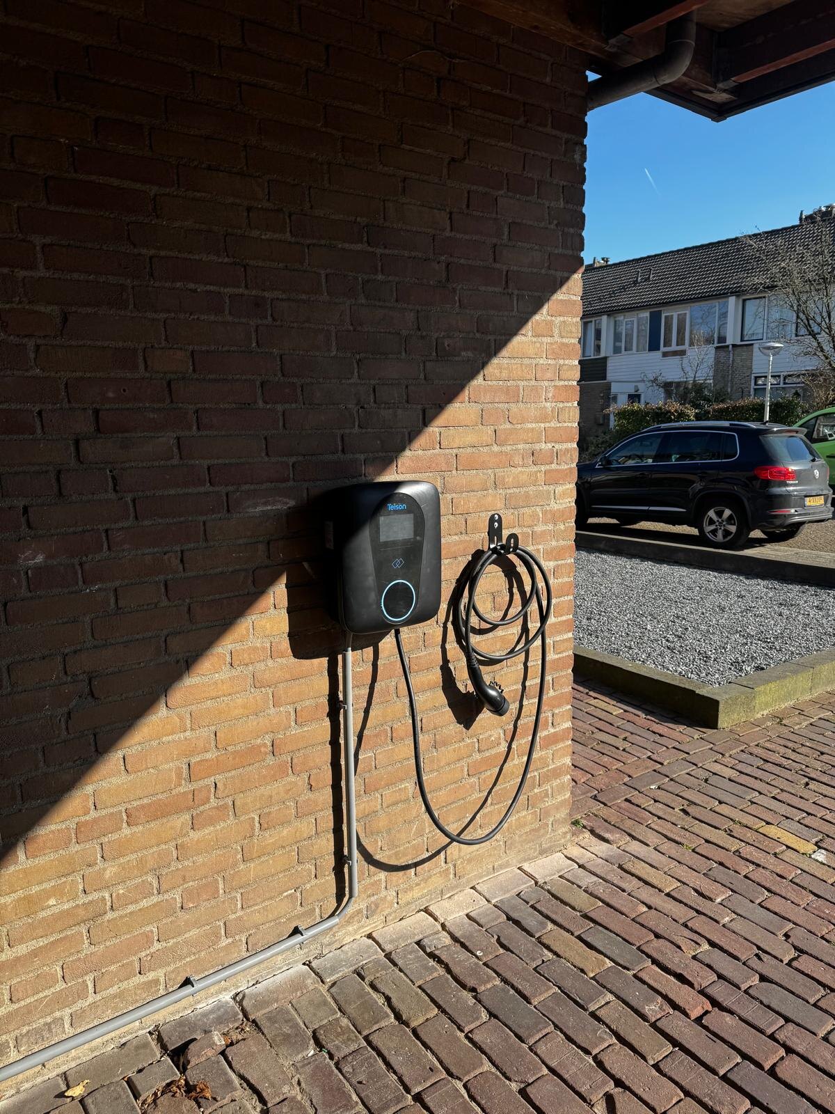 Laadpaal aan de gevel van een woning met opgerolde laadkabel en auto op de oprit