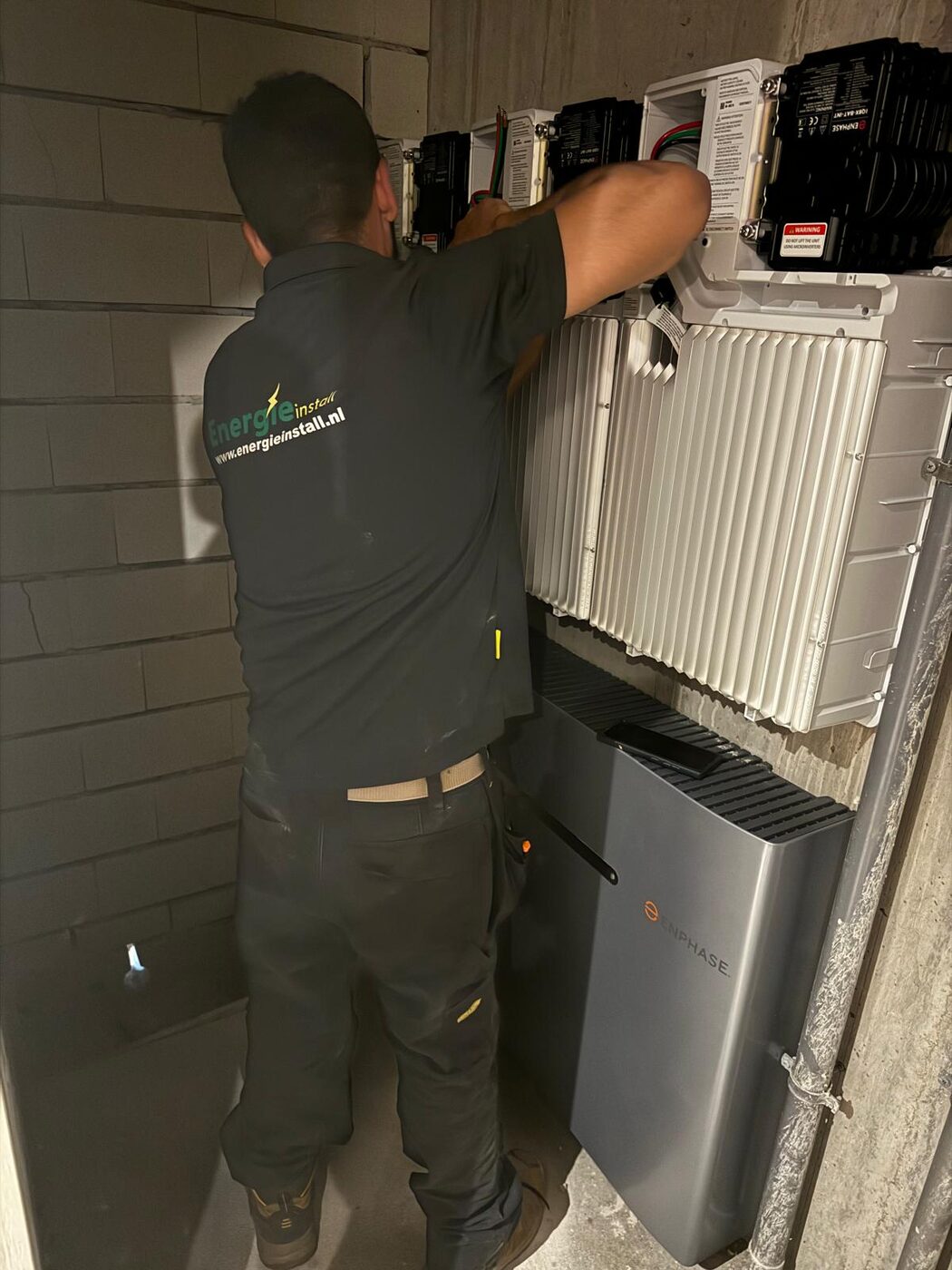 Monteur van Energie Install in bedrijfskleding sluit een omvormer aan boven een Enphase thuisbatterij