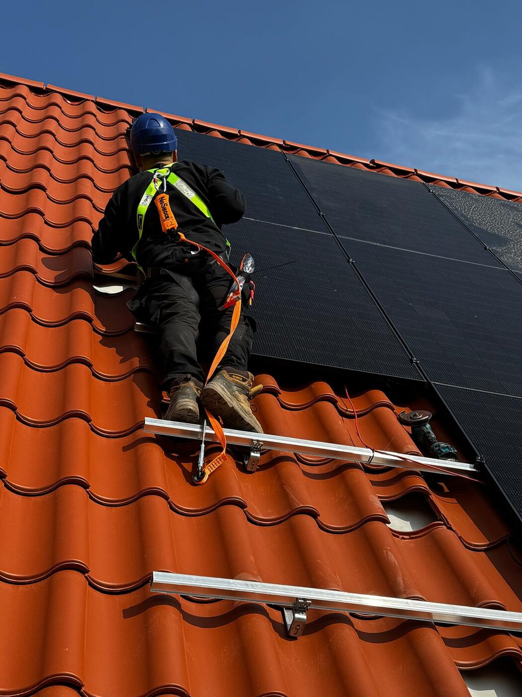 Monteur met veiligheidsharnas en helm installeert zwarte zonnepanelen op een rood pannendak met montagerails
