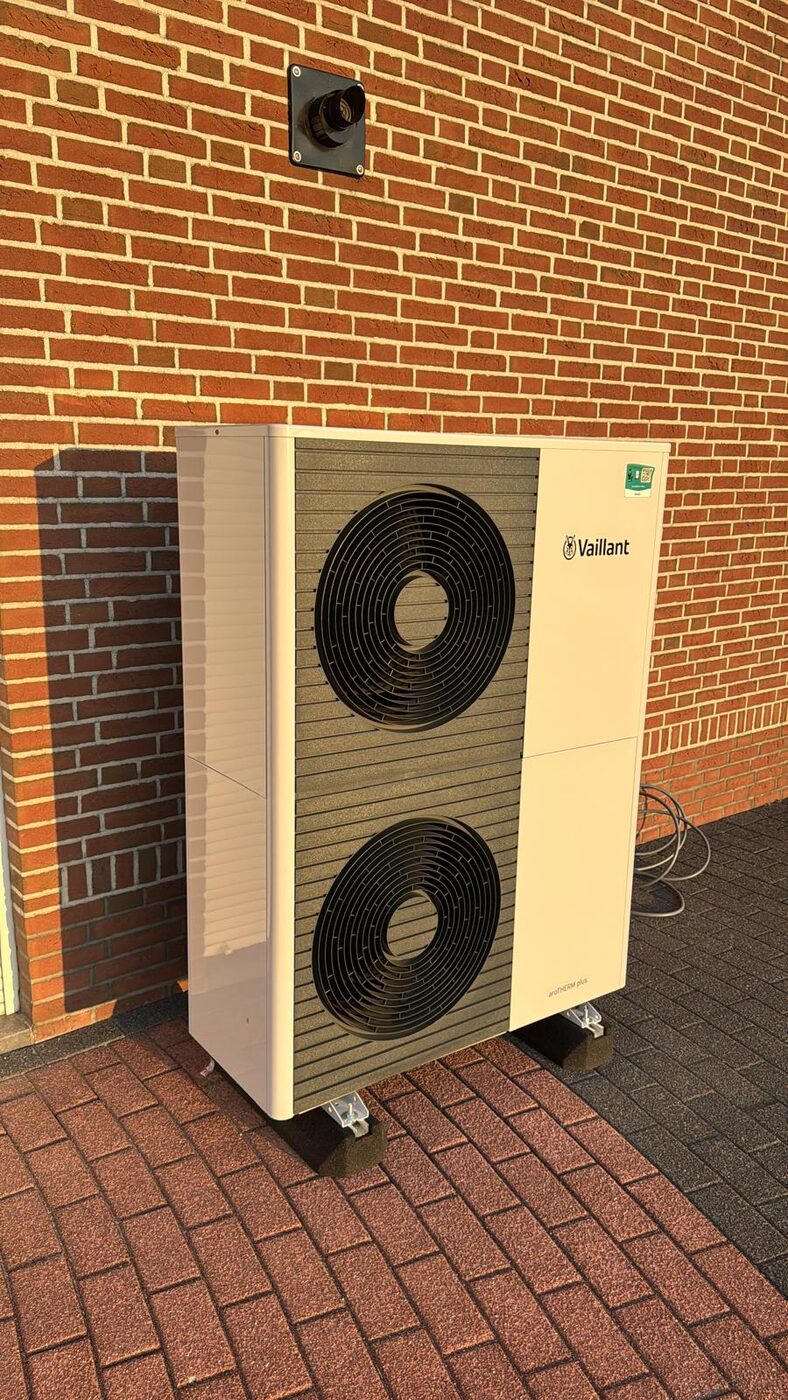 Vaillant lucht-water warmtepomp met dubbele ventilator tegen bakstenen gevel op klinkerterras in warm avondlicht