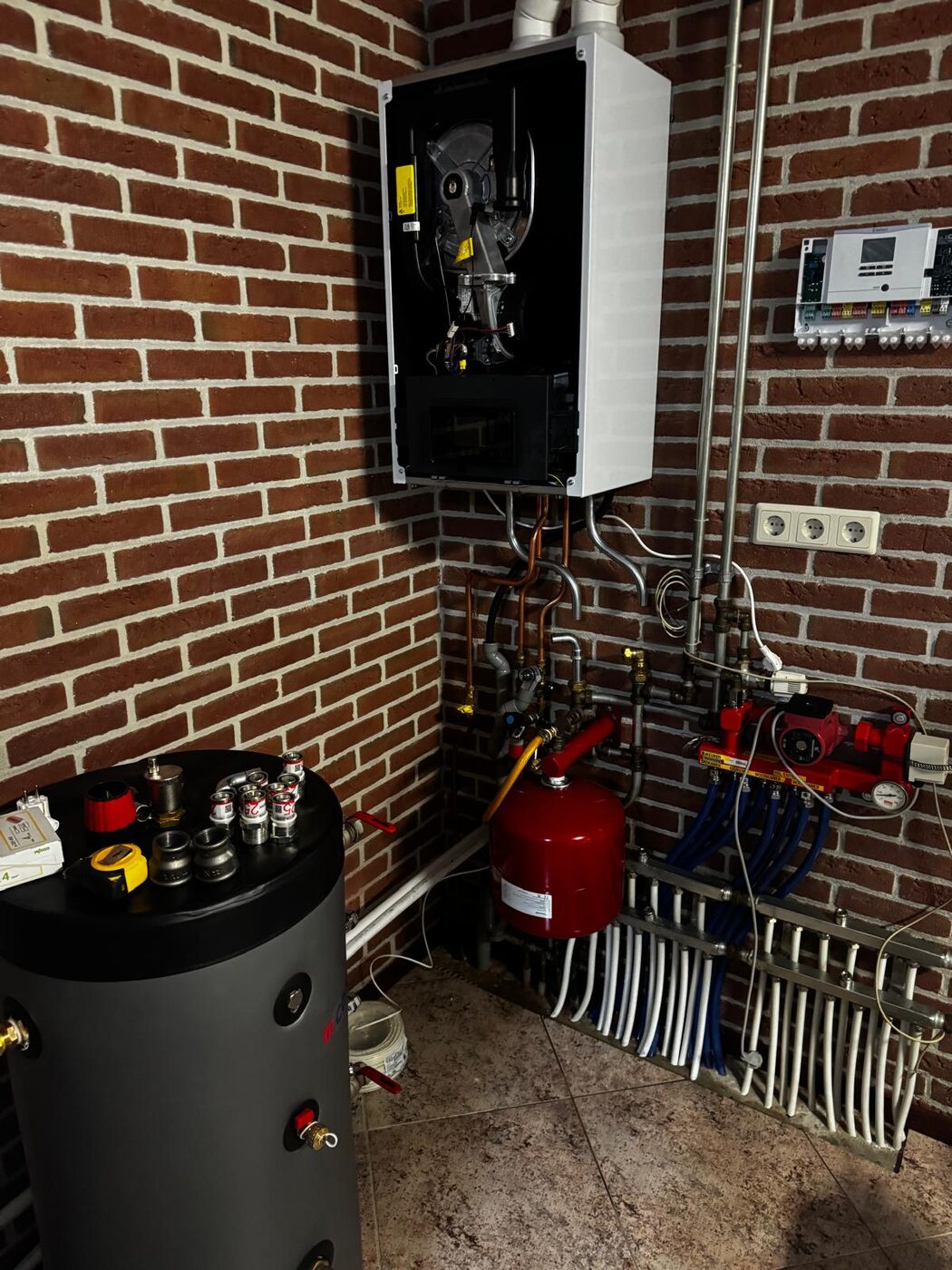 Binnenopstelling van een warmtepompinstallatie met buffervat, leidingwerk en regeltechniek in een technische ruimte tegen een bakstenen muur