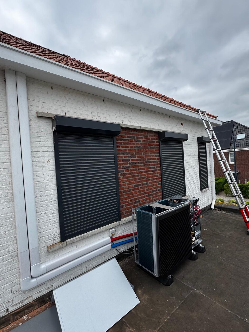 Buitenunit van een lucht-water warmtepomp geinstalleerd tegen de gevel van een woning met rolluiken