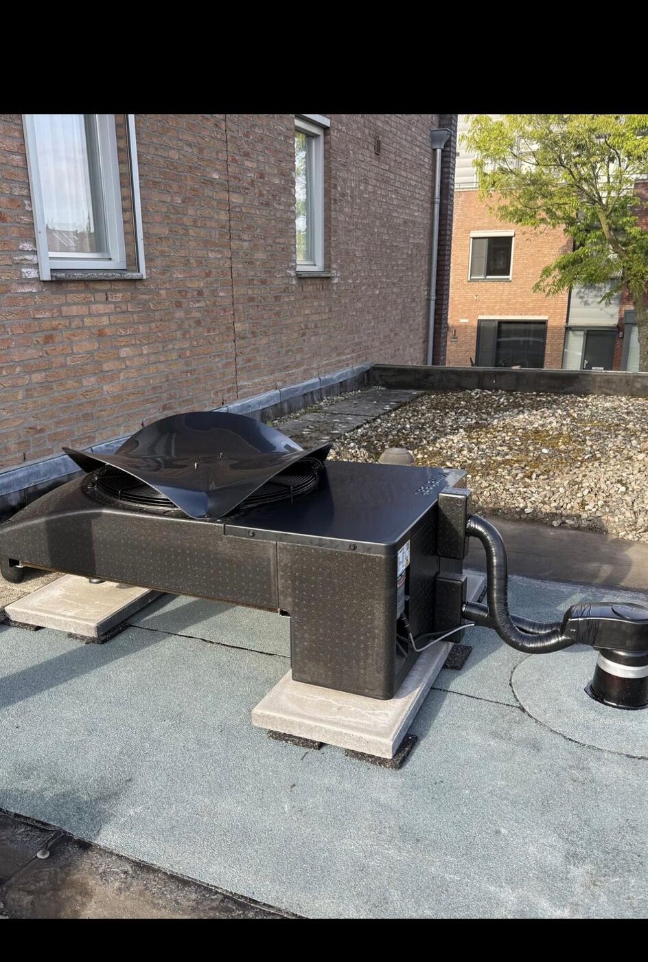 Zwarte warmtepomp buitenunit op stelpoten op plat dak met grind naast bakstenen woning