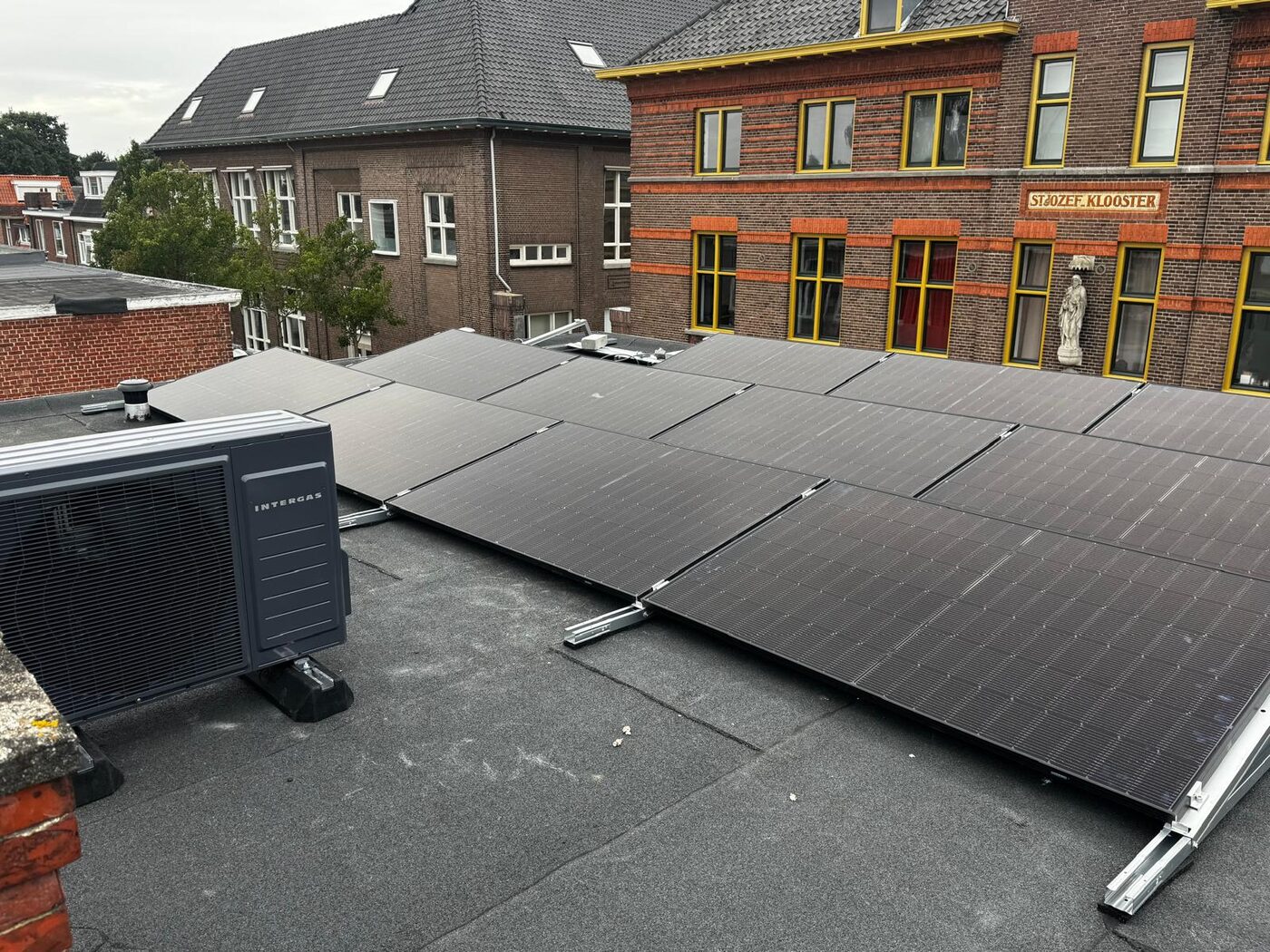 Zonnepanelen en een buitenunit van een warmtepomp op een plat dak tegenover het St. Jozef Klooster