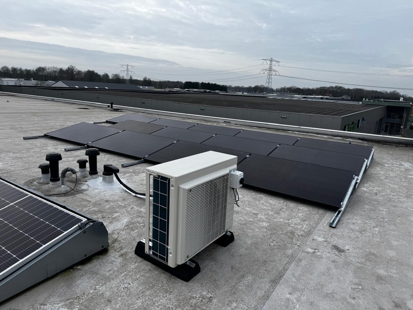 Zonnepanelen en warmtepomp-buitendeel op plat bedrijfsdak met hoogspanningsmasten op de achtergrond, installatie door Energie Install.
